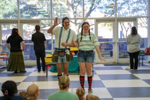 Two smiling performers stand arm-in-arm in front of an audience in a bright indoor space. One waves while wearing a utility belt and shorts; the other wears a striped shirt, shorts, colorful knee-high socks, and braided hair. Several people stand near tables in the background.