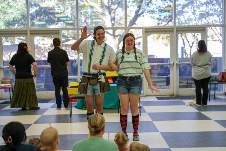 Two smiling performers stand arm-in-arm in front of an audience in a bright indoor space. One waves while wearing a utility belt and shorts; the other wears a striped shirt, shorts, colorful knee-high socks, and braided hair. Several people stand near tables in the background.