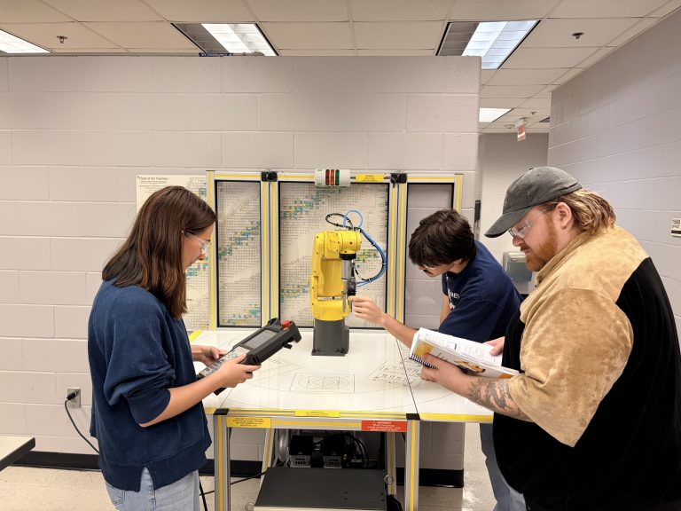 Three people wearing safety glasses work around a yellow industrial robotic arm in a laboratory. One person operates a handheld controller, another adjusts the robot, and a third consults an instruction manual.