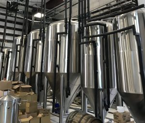 Interior of an industrial facility with rows of large stainless-steel tanks connected by an extensive network of black insulated pipes suspended overhead and running vertically between the tanks.