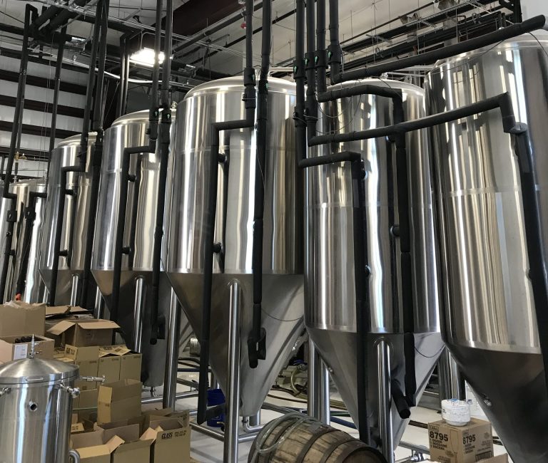 Interior of an industrial facility with rows of large stainless-steel tanks connected by an extensive network of black insulated pipes suspended overhead and running vertically between the tanks.