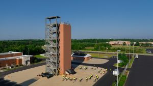 Aerial view of a multi-story fire training tower with exterior metal staircases at a fire station. Firefighters in bright yellow shirts participate in outdoor training exercises around the tower on a sunny day.
