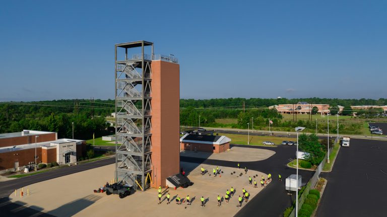 Aerial view of a multi-story fire training tower with exterior metal staircases at a fire station. Firefighters in bright yellow shirts participate in outdoor training exercises around the tower on a sunny day.