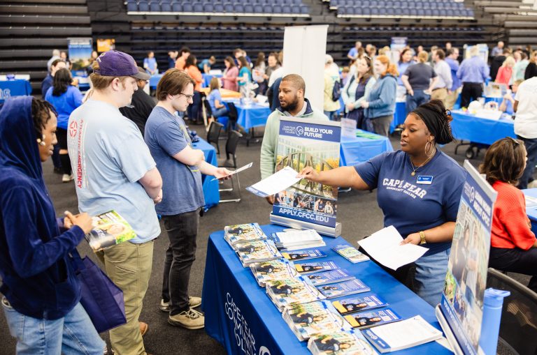 A Cape Fear Community College representative hands an informational flyer to attendees at a busy college fair. Brochures and promotional materials cover a blue table, while visitors explore other booths in the background.
