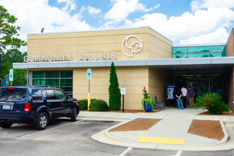 Exterior of a tan brick community college building with a large college logo and Alexander Guy Building signage. A blue SUV is parked near the entrance, where several people stand by the glass doors.