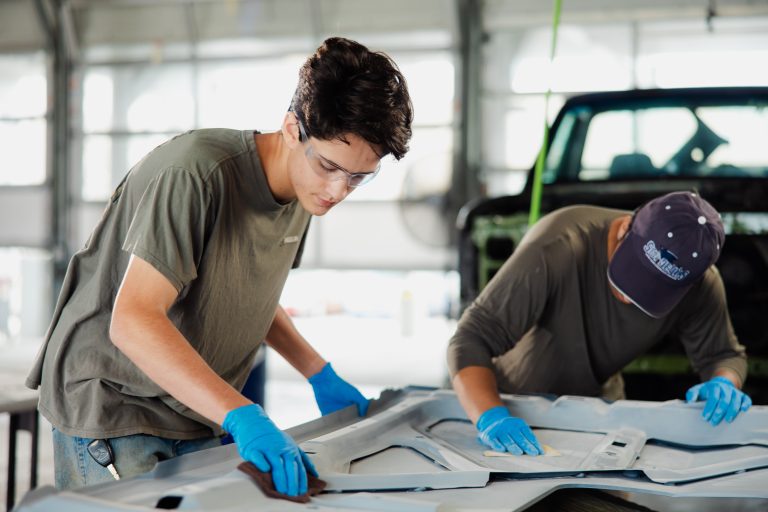 Two automotive technicians wearing blue gloves sand and prepare a vehicle body panel in a workshop.
