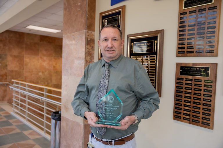 A man wearing a green dress shirt and patterned tie holds a glass Partnership Award recognizing Timothy L. Fuss of Cape Fear Community College at the 2026 Basic Law Enforcement Training Directors’ Conference. Framed law enforcement leadership award plaques hang on the wall behind him.