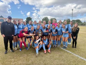 CFCC's women's soccer team in light blue uniforms celebrates on the field, smiling and posing with an NJCAA Region 10 championship plaque. Coaches and staff join the team for the group photo.