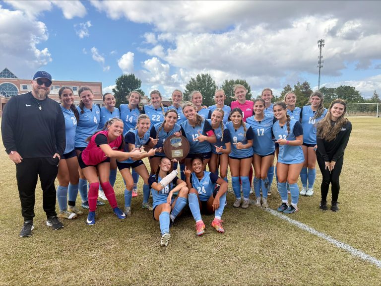 CFCC's women's soccer team in light blue uniforms celebrates on the field, smiling and posing with an NJCAA Region 10 championship plaque. Coaches and staff join the team for the group photo.