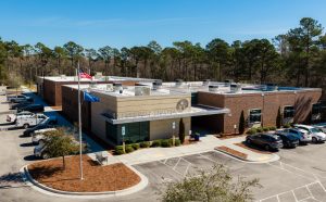 Aerial view of Cape Fear Community College's brick campus building, with American and college flags, surrounding parking lots, and a backdrop of tall pine trees under a clear blue sky.