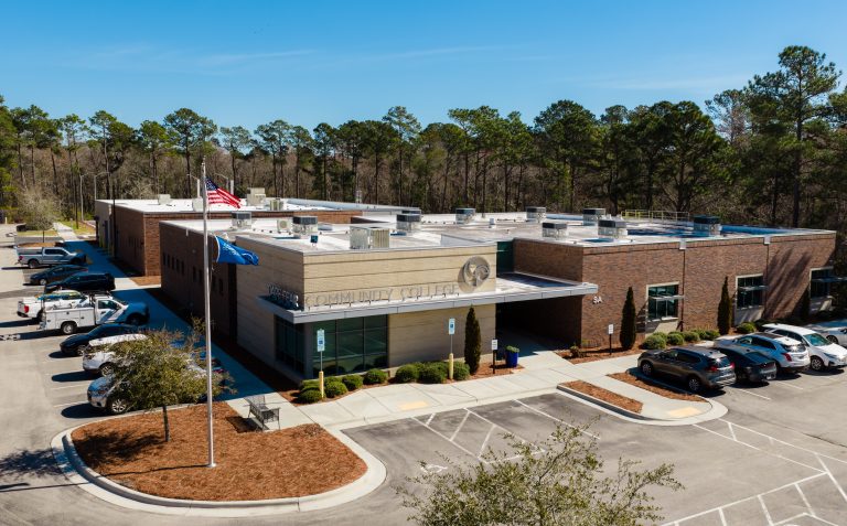 Aerial view of Cape Fear Community College's brick campus building, with American and college flags, surrounding parking lots, and a backdrop of tall pine trees under a clear blue sky.
