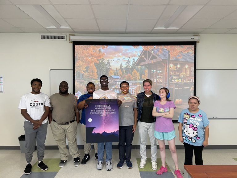 Eight people stand together in a classroom in front of a projected autumn café scene. Two people in the center hold a purple poster featuring a moonlit mountain landscape and text about dancing under the stars.