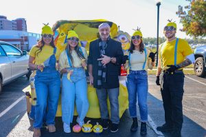 Five adults pose in a parking lot, four dressed as Minions in yellow shirts, blue overalls, yellow hats, and goggles, alongside a person dressed as Gru in a black coat and gray scarf. Behind them is a Minions-themed decorated vehicle, with painted pumpkins on the ground.