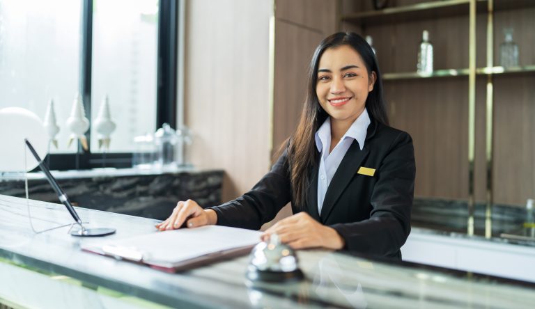 Smiling female receptionist wearing a black blazer and white collared shirt, seated behind a marble reception desk with a guest register and service bell in a modern hotel lobby.
